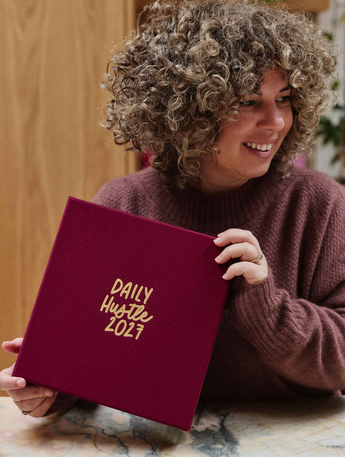 A smiling woman with curly hair holds a plum hardcover planner with gold foil text reading 'DAILY HUSTLE 2027' on the cover, set against a warm indoor background.