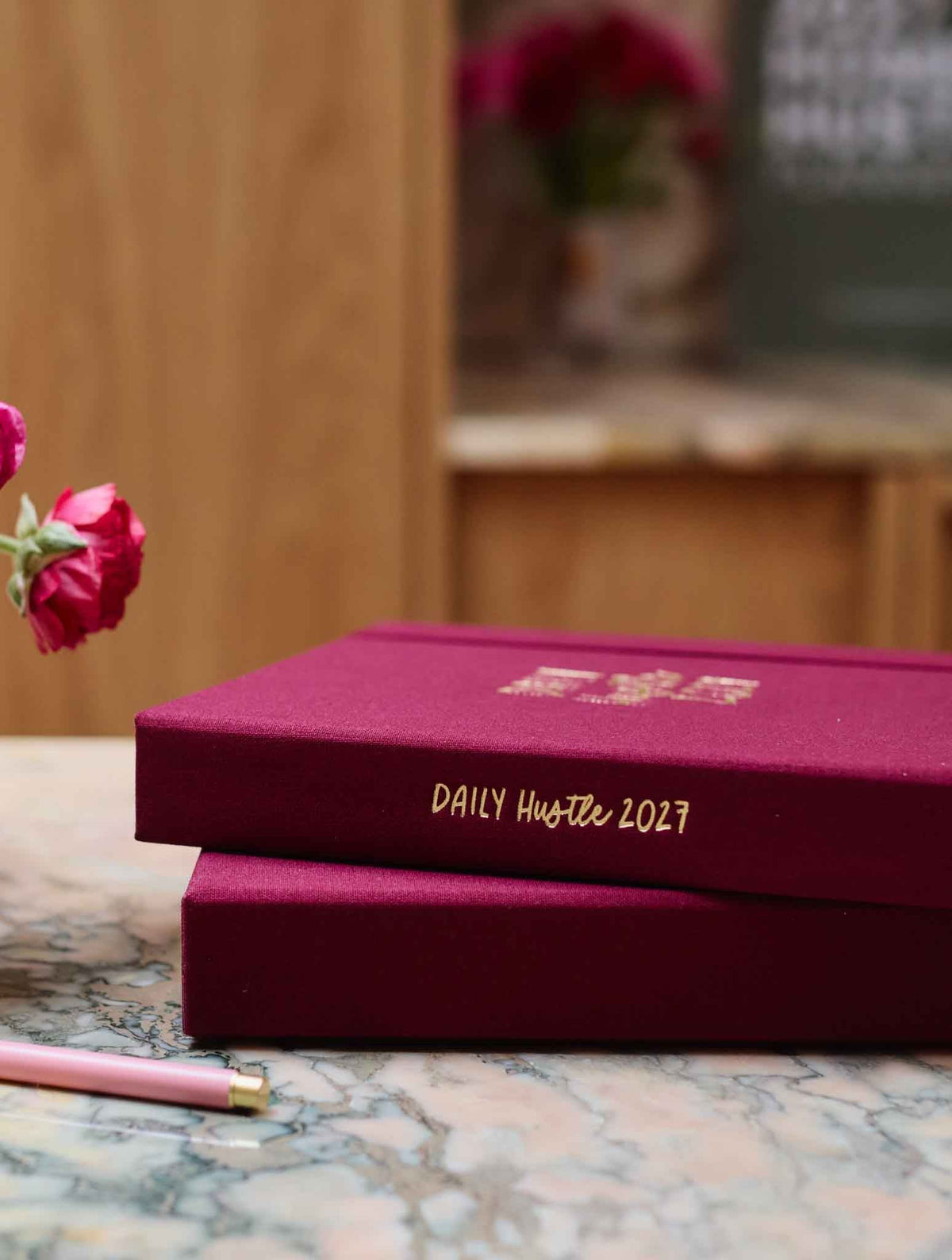 A hardcover planner in plum stacked on its box with gold foil lettering reading 'DAILY Hustle 2027' on a marble surface, with a blurred wooden interior and pink roses in the background.