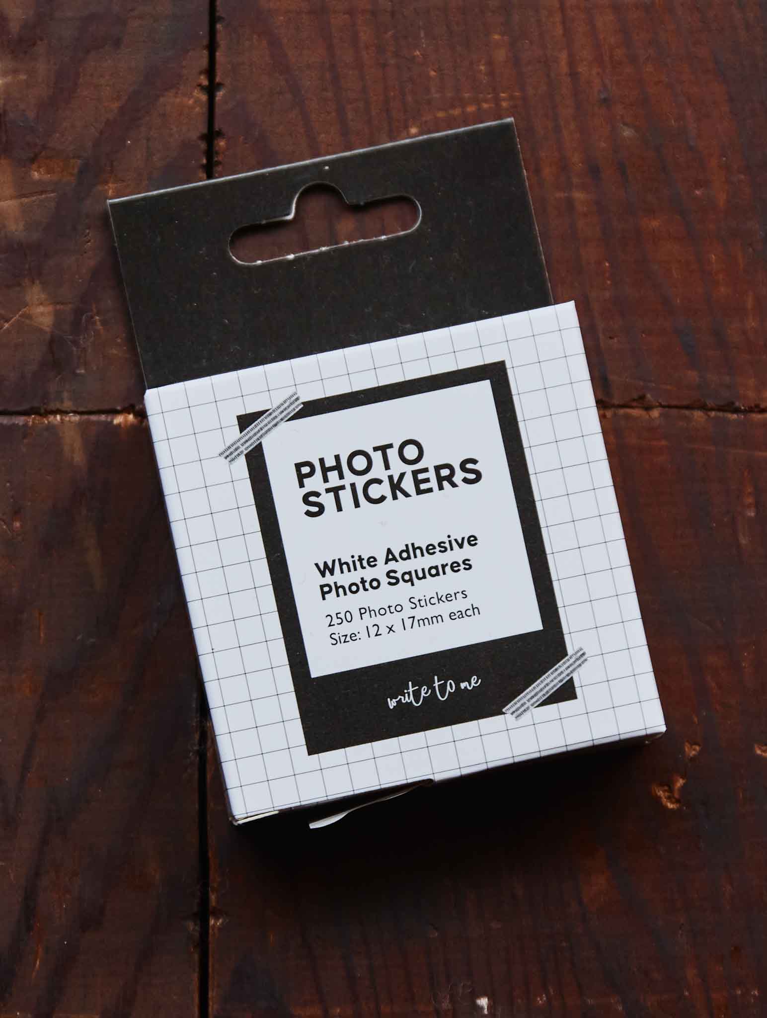 A package of white adhesive photo stickers in a black and white grid-patterned box, photographed on a dark wooden surface.
