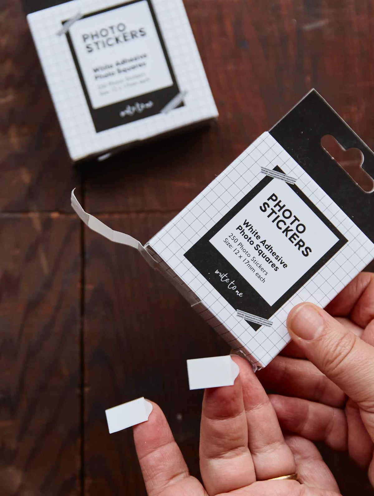 A person's hands hold a package of white adhesive photo stickers over a dark wooden table, with another box blurred in the background.