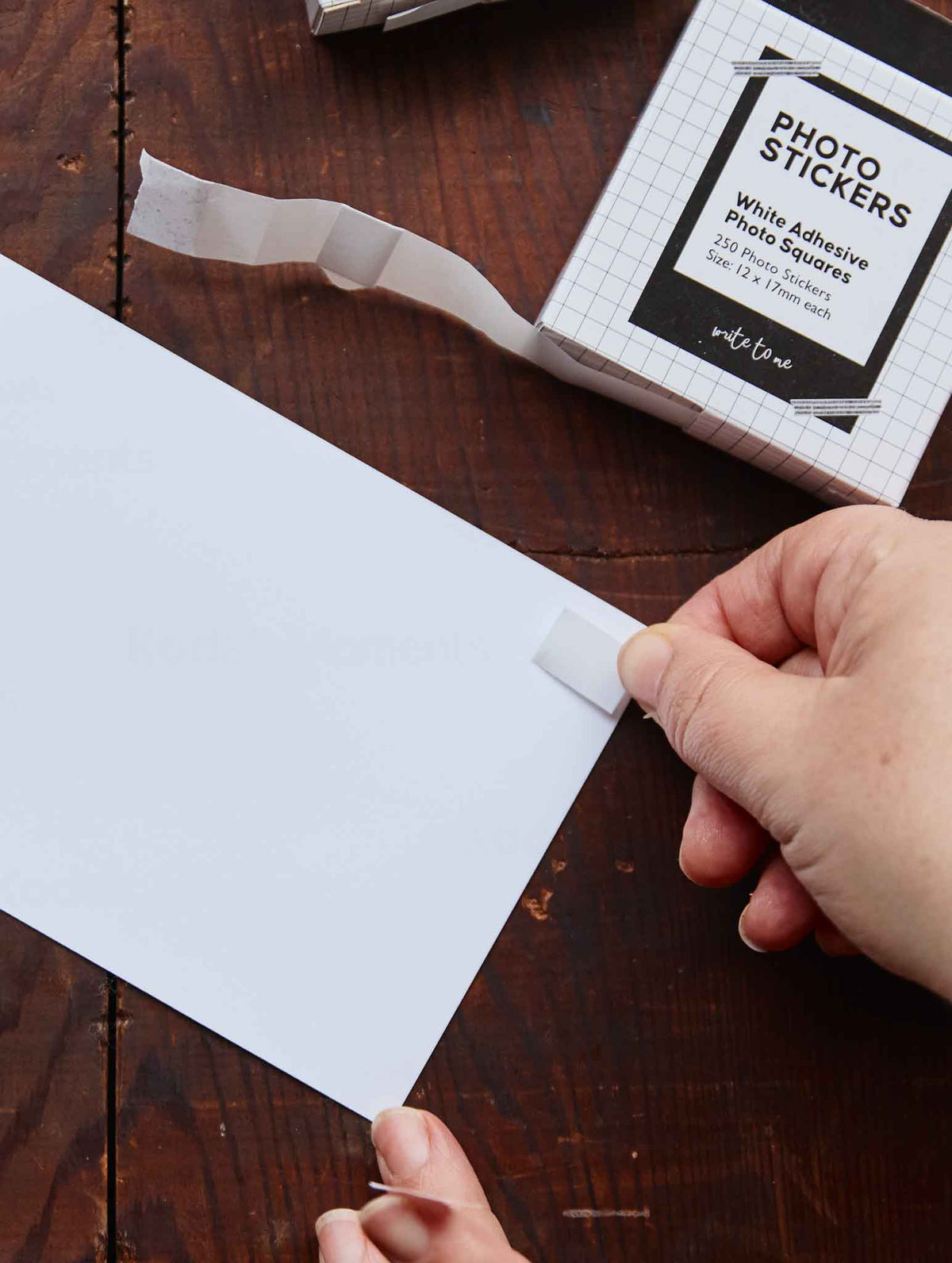 A person's hands peel a white adhesive photo sticker from its grid-patterned packaging box and puts it on a photo, set on a dark wooden tabletop.