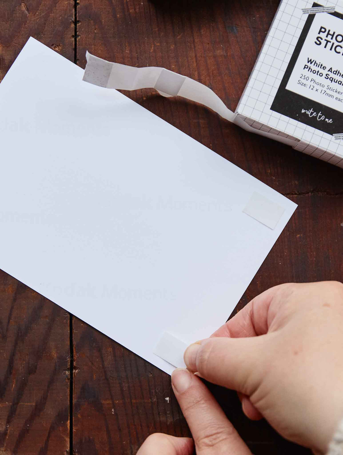 A person's hands are shown applying a small square of white adhesive tape to the corner of a photo, with a roll of white photo stickers on a dark wooden table.