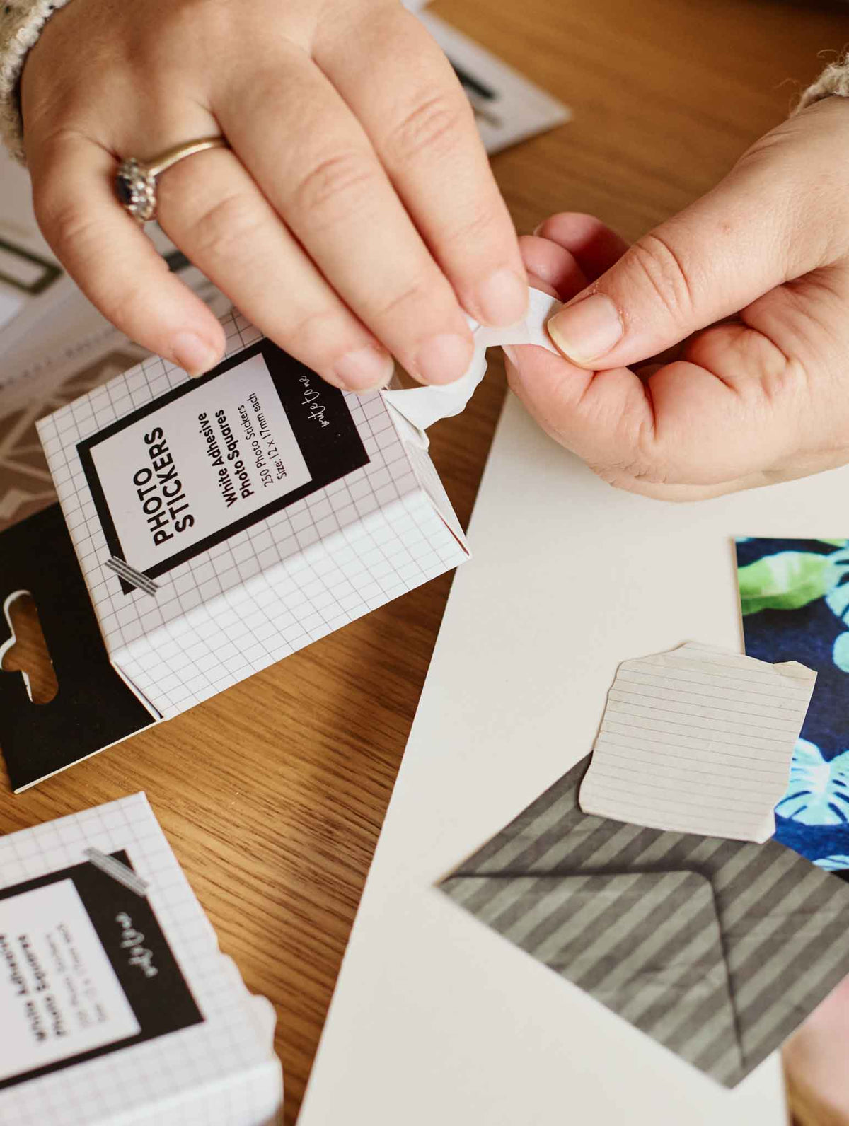 A close-up of hands peeling a small white photo sticker from its grid-patterned packaging box on a wooden table.
