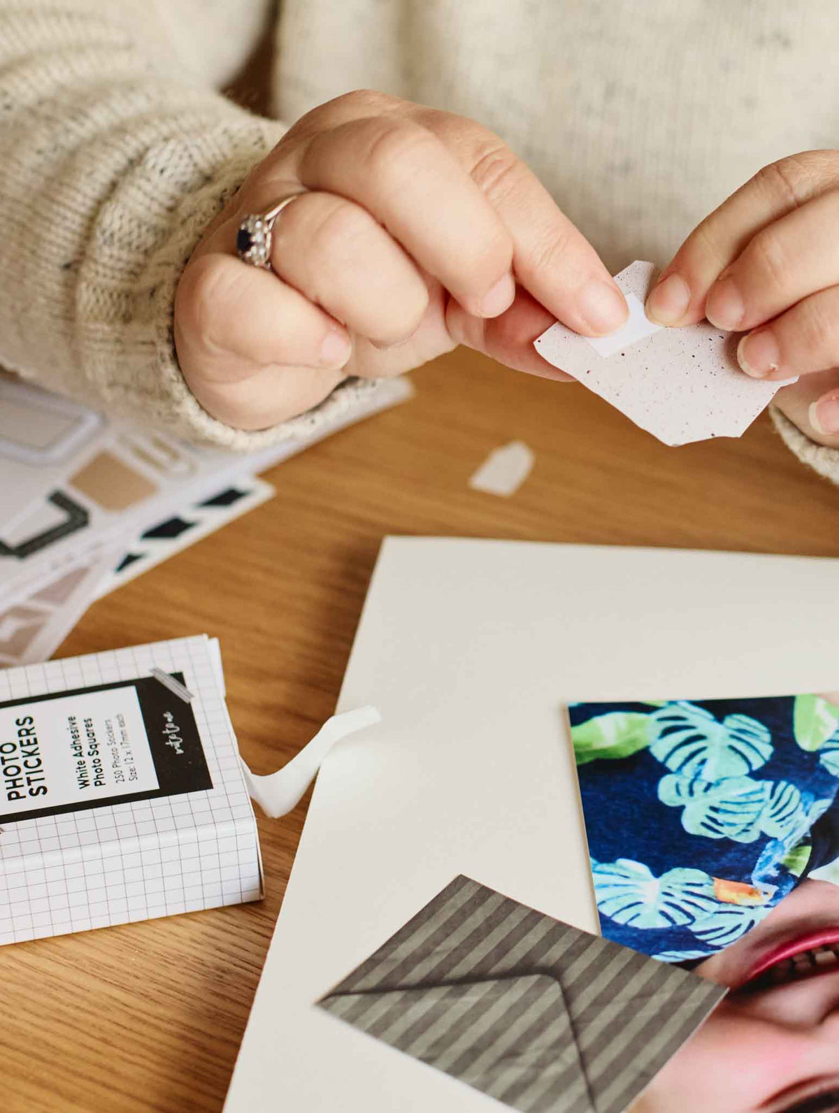 A close-up of a person's hands holding a small piece of textured white paper over a wooden desk, surrounded by craft supplies including a box of photo stickers, a colorful tropical print, and a gray striped envelope.